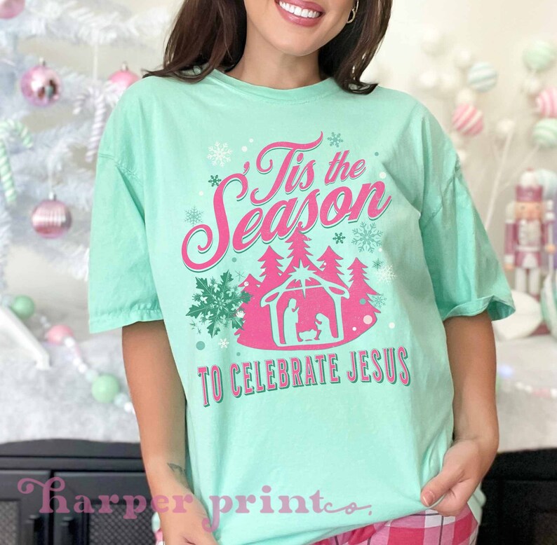 May include: A light green t-shirt with a pink and green Christmas design. The design features the text "Tis the Season to Celebrate Jesus" with a pink nativity scene and snowflakes.