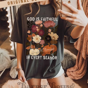 May include: Dark gray t-shirt with a floral and pumpkin graphic. The design includes the text "GOD IS FAITHFUL" and "IN EVERY SEASON". The shirt also features the text "harper house THREE SIXTEEN" at the bottom.