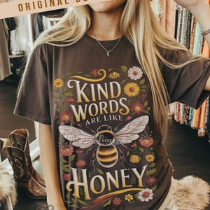 Peut inclure: T-shirt marron fonc&eacute; avec l'inscription "KIND WORDS ARE LIKE HONEY" dans une police d&eacute;corative, avec une illustration d'abeille entour&eacute;e de fleurs color&eacute;es. Le t-shirt a des manches courtes et une coupe d&eacute;contract&eacute;e.