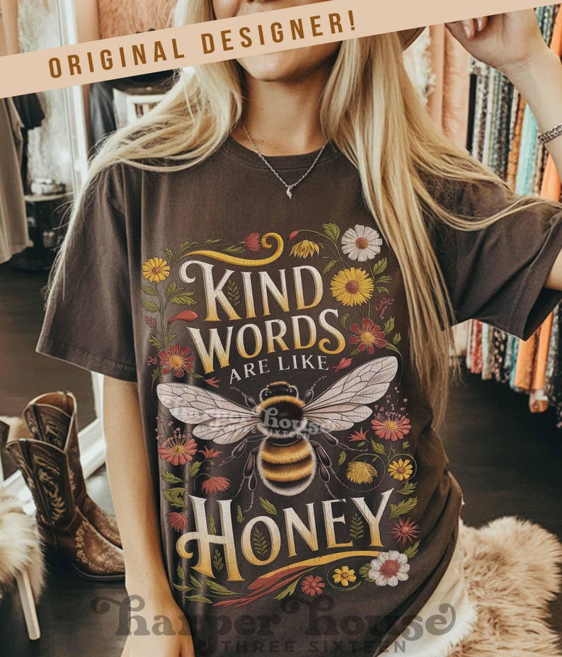 Peut inclure: T-shirt marron fonc&eacute; avec l'inscription "KIND WORDS ARE LIKE HONEY" dans une police d&eacute;corative, avec une illustration d'abeille entour&eacute;e de fleurs color&eacute;es. Le t-shirt a des manches courtes et une coupe d&eacute;contract&eacute;e.