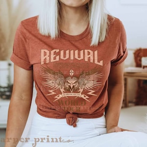 May include: A rust-colored t-shirt with a graphic design of an eagle with wings spread, a cross, and the text "REVIVAL 2 CHRONICLES 7:14 WORLD TOUR".
