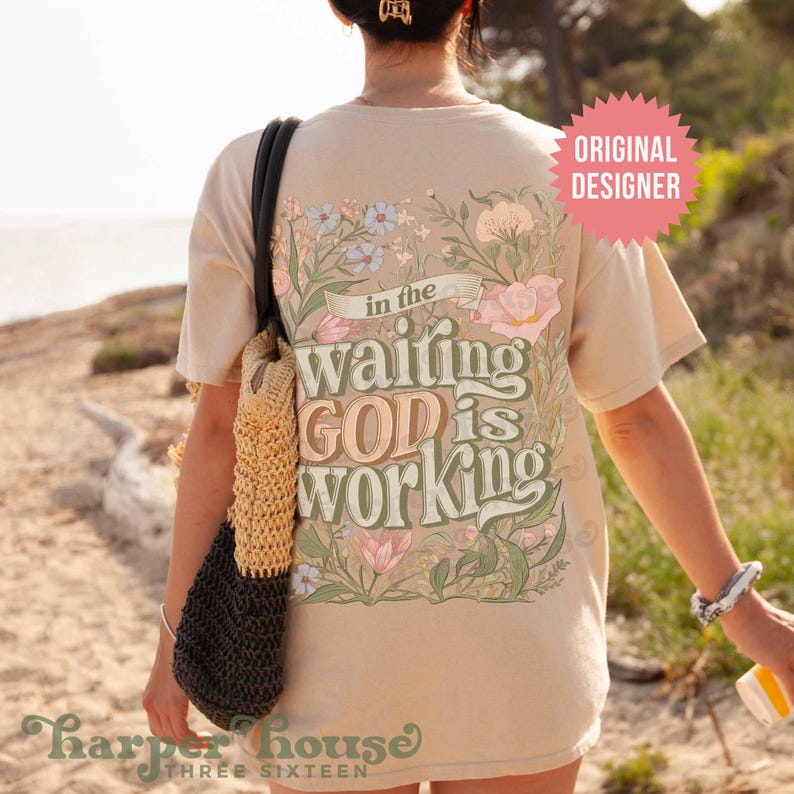 May include: A beige t-shirt with a floral design and the text "in the waiting GOD is working" printed on the back. The shirt is being worn by a person.