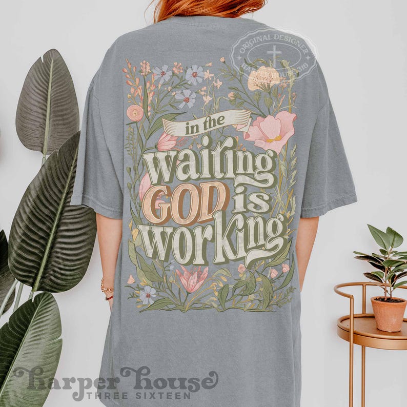 May include: A light grey t-shirt with a floral design and the text "In the waiting GOD is working".