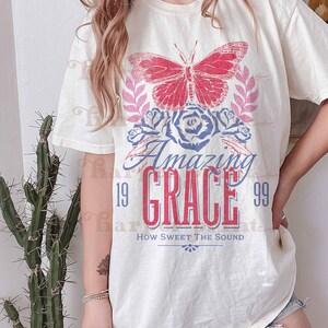 Pu&ograve; includere: Una maglietta bianca con una grafica di farfalla rossa e blu, rose e il testo "Amazing Grace 19 99 How Sweet The Sound".