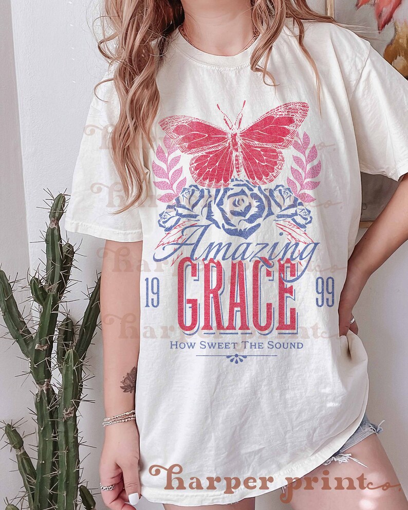 Pu&ograve; includere: Una maglietta bianca con una grafica di farfalla rossa e blu, rose e il testo "Amazing Grace 19 99 How Sweet The Sound".
