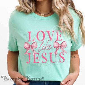 K&ouml;nnte beinhalten: Hellgr&uuml;nes T-Shirt mit dem pinkfarbenen Schriftzug "LOVE like JESUS" und zwei rosa Schleifen. Das T-Shirt hat einen Rundhalsausschnitt und kurze &Auml;rmel. Der Markenname "Harper House" ist unten aufgedruckt.