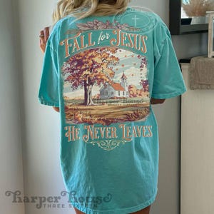 May include: A turquoise t-shirt with the words "FALL for JESUS" and "HE NEVER LEAVES" in an orange script. The back of the shirt features a graphic of a church, a tree with autumn leaves, and the text "Harper House Three Sixteen."