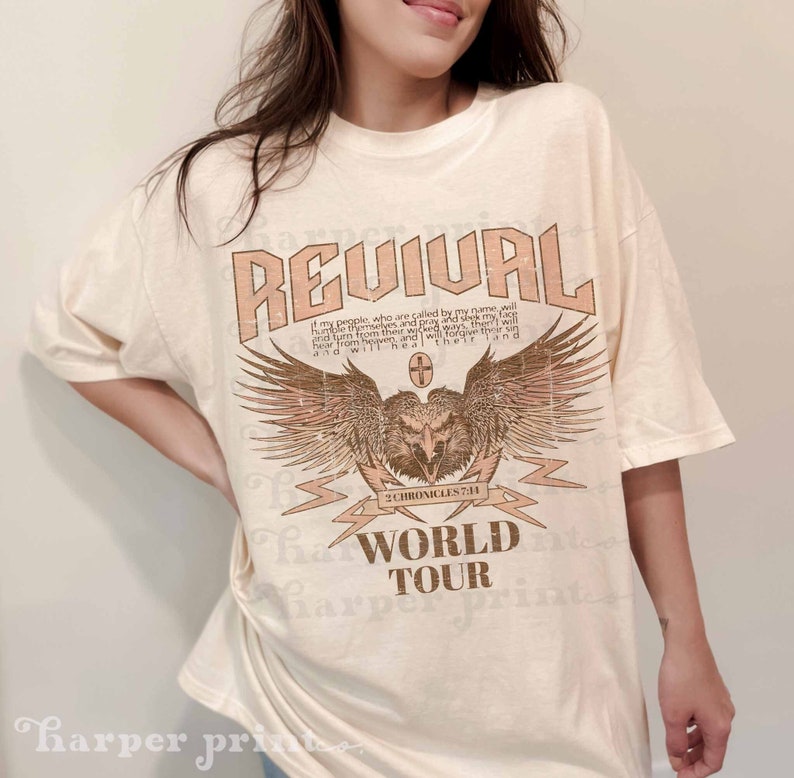 May include: A white t-shirt with a distressed graphic design featuring an eagle with wings spread, a cross, and the text "REVIVAL", "2 CHRONICLES 7:11", "WORLD TOUR".