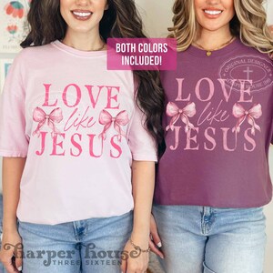 K&ouml;nnte beinhalten: Zwei T-Shirts, eines hellrosa und eines lila, mit dem Text "LOVE like JESUS" in Rosa. Das Design beinhaltet zwei rosa Schleifen. Der Satz "BOTH COLORS INCLUDED!" befindet sich in einem rosa Rechteck oben.