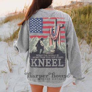 Pode incluir: Uma camisa de moletom cinza com uma estampa gr&aacute;fica de um soldado ajoelhado em frente a uma cruz coberta com uma bandeira americana. O texto "When the world is more than you can stand: Kneel" est&aacute; impresso abaixo do gr&aacute;fico. O texto "Harper House Three Sixteen" est&aacute; impresso na parte inferior da camisa de moletom.