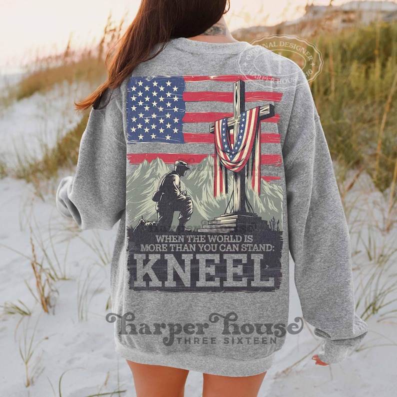 Pode incluir: Uma camisa de moletom cinza com uma estampa gr&aacute;fica de um soldado ajoelhado em frente a uma cruz coberta com uma bandeira americana. O texto "When the world is more than you can stand: Kneel" est&aacute; impresso abaixo do gr&aacute;fico. O texto "Harper House Three Sixteen" est&aacute; impresso na parte inferior da camisa de moletom.