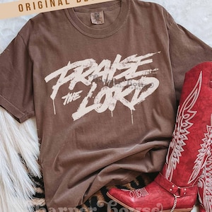 Può includere: Una maglietta marrone con le parole "PRAISE THE LORD" in un carattere bianco invecchiato. La maglietta è abbinata a stivali da cowboy rossi con cuciture bianche. Gli stivali hanno un piccolo tacco e un dettaglio a catena.