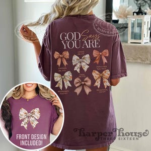 Op de afbeelding: Een donker mauve t-shirt met korte mouwen en een achteropdruk met de tekst GOD Says YOU ARE en zes geïllustreerde strikken met bijbelverwijzingen. De voorkant van het t-shirt toont een enkele bloemrijke strik met de tekst FRONT DESIGN INCLUDED.