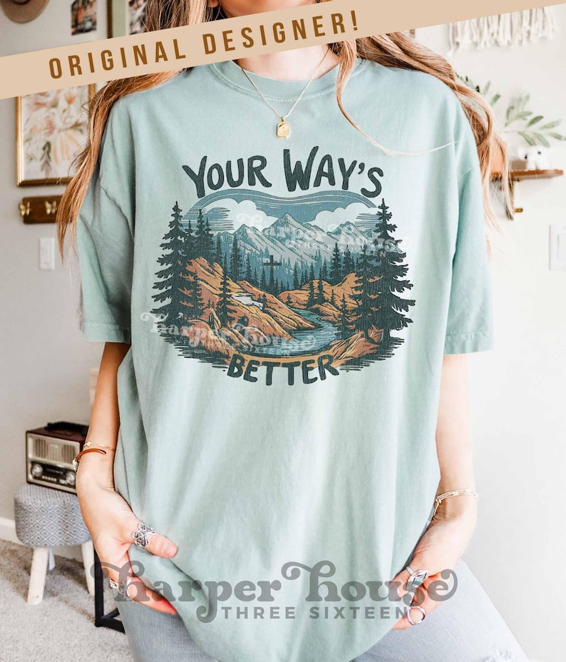 May include: A light green t-shirt with a graphic of a mountain landscape, river, and trees. The text "YOUR WAY'S BETTER" is above the landscape. The words "ORIGINAL DESIGNER!" are printed on a banner at the top.