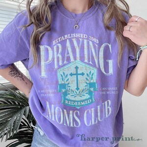 Op de afbeelding: Paars T-shirt met een vintage-stijl grafisch ontwerp. De grafiek toont een schild met een kruis, de woorden "Praying Moms Club" en de tekst "Established 2003".