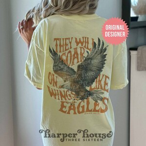 May include: A yellow t-shirt with a vintage-style graphic of an eagle with the text "They Will Soar On Wings Like Eagles Isaiah 40:31" printed on the back. The shirt also has the text "Harper House Three Sixteen" printed on the bottom.