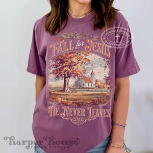 May include: A purple t-shirt with the text "Fall for Jesus" and "He Never Leaves" in an ornate design. The graphic features a church and a tree with fall foliage. The shirt has a relaxed fit.