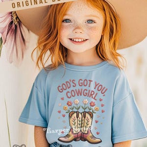 Op de afbeelding: Een lichtblauw T-shirt met de tekst "GOD'S GOT YOU COWGIRL" in rode borduursels. Het shirt heeft een ontwerp van cowboylaarzen met bloemen. De woorden "ORIGINAL DESIGNER!" staan bovenaan.