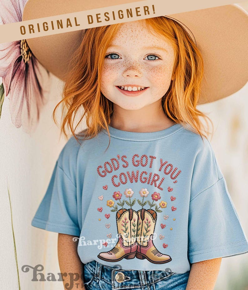 Op de afbeelding: Een lichtblauw T-shirt met de tekst "GOD'S GOT YOU COWGIRL" in rode borduursels. Het shirt heeft een ontwerp van cowboylaarzen met bloemen. De woorden "ORIGINAL DESIGNER!" staan bovenaan.