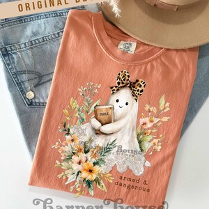May include: Peach-colored t-shirt featuring a cartoon ghost holding a Bible, adorned with a leopard print bow and surrounded by floral illustrations. The text "armed & dangerous" is below the ghost. The shirt is folded on top of blue jeans.