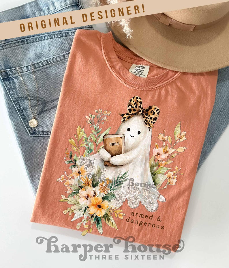 May include: Peach-colored t-shirt featuring a cartoon ghost holding a Bible, adorned with a leopard print bow and surrounded by floral illustrations. The text "armed & dangerous" is below the ghost. The shirt is folded on top of blue jeans.