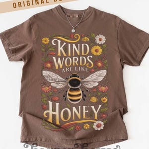 Peut inclure: T-shirt marron avec l'inscription "KIND WORDS ARE LIKE HONEY" et une illustration d'abeille. Le motif est entour&eacute; de fleurs color&eacute;es. Le t-shirt porte &eacute;galement l'inscription "ORIGINAL DESIGNER!" en haut et "harper house THREE SIXTEEN" en bas.