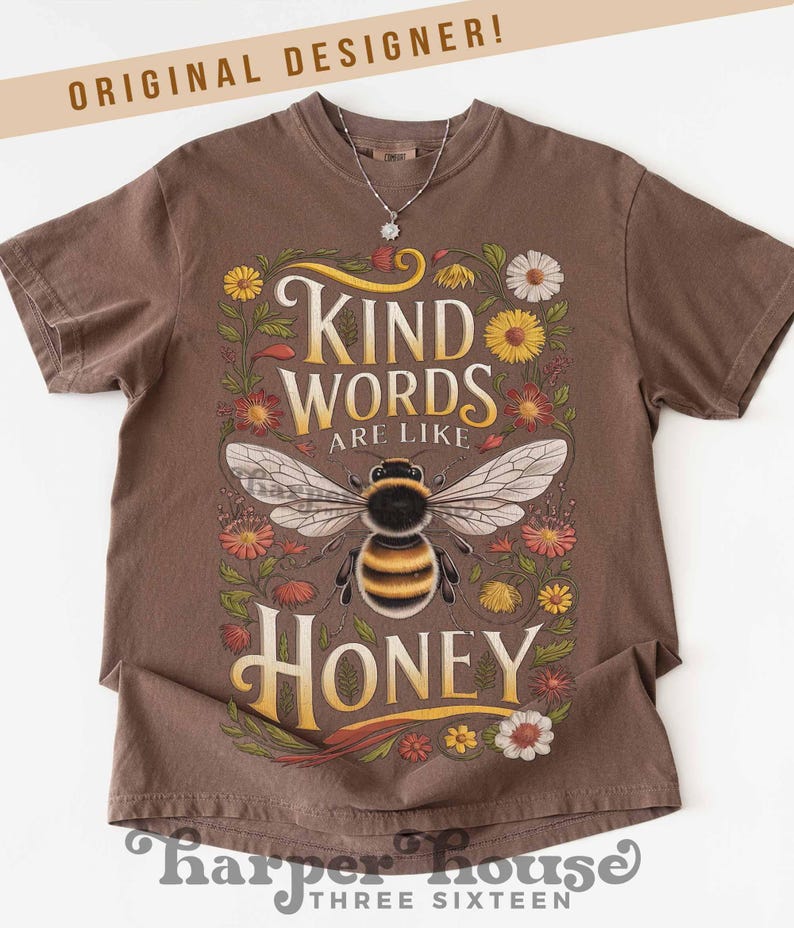 Peut inclure: T-shirt marron avec l'inscription "KIND WORDS ARE LIKE HONEY" et une illustration d'abeille. Le motif est entour&eacute; de fleurs color&eacute;es. Le t-shirt porte &eacute;galement l'inscription "ORIGINAL DESIGNER!" en haut et "harper house THREE SIXTEEN" en bas.