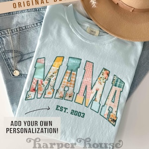 May include: Light blue t-shirt with a patchwork design that spells out "MAMA" in large letters. The letters are outlined in yellow and have a floral and plaid pattern. The text "EST. 2003" is printed below the letters.