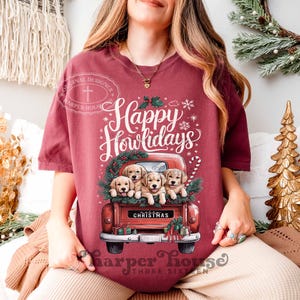 Op de afbeelding: Een maroon t-shirt met een rode vrachtwagen vol met golden retriever puppy's. De vrachtwagen heeft een "Christmas" bord aan de achterkant. De tekst "Happy Howlidays" staat boven de vrachtwagen.