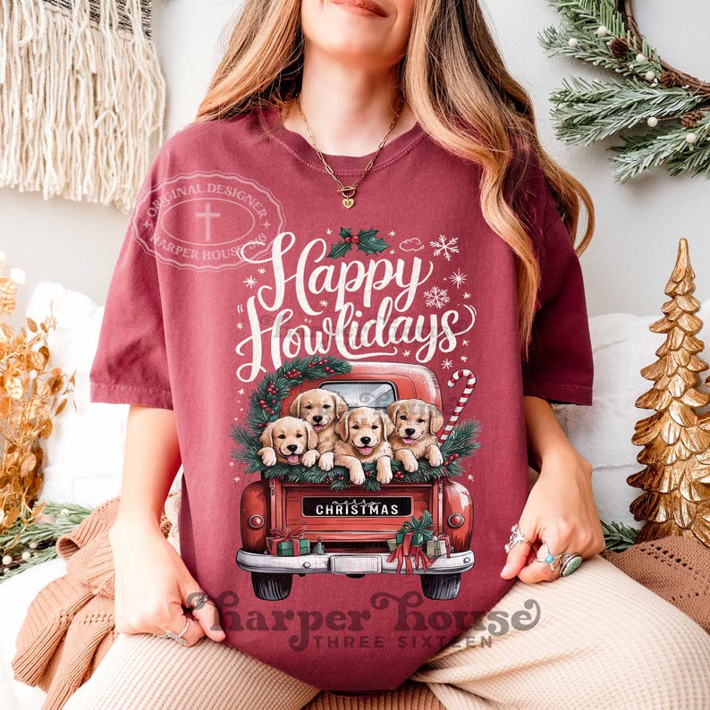 Op de afbeelding: Een maroon t-shirt met een rode vrachtwagen vol met golden retriever puppy's. De vrachtwagen heeft een "Christmas" bord aan de achterkant. De tekst "Happy Howlidays" staat boven de vrachtwagen.