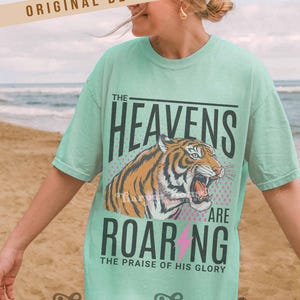 May include: A light green t-shirt with the text "THE HEAVENS ARE ROARING" and a tiger graphic. The shirt also includes the text "THE PRAISE OF HIS GLORY" and the brand name "harper house THREE SIXTEEN".