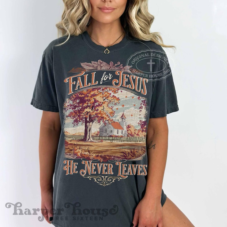 May include: Dark gray t-shirt with the text "Fall for Jesus" above a fall landscape scene with a church and tree. The text "He Never Leaves" is below the image. The shirt has a vintage design.