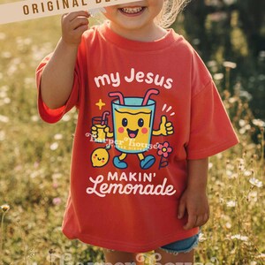 Op de afbeelding: Rood t-shirt met de witte tekst "my Jesus makin' lemonade". Het shirt heeft een cartoonafbeelding van een glimlachend limonadeglas met armen en benen, dat een duim omhoog geeft. Er is ook een klein citroenpersonage aanwezig.