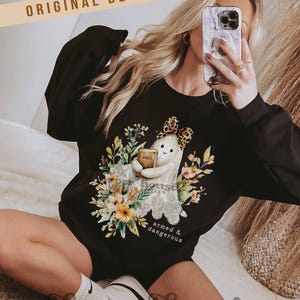 May include: Black sweatshirt featuring a ghost design holding a book labeled "Bible" surrounded by floral elements. The ghost wears a leopard print bow. Text on the sweatshirt reads "armed & dangerous". The words "Original Designer!" are at the top.