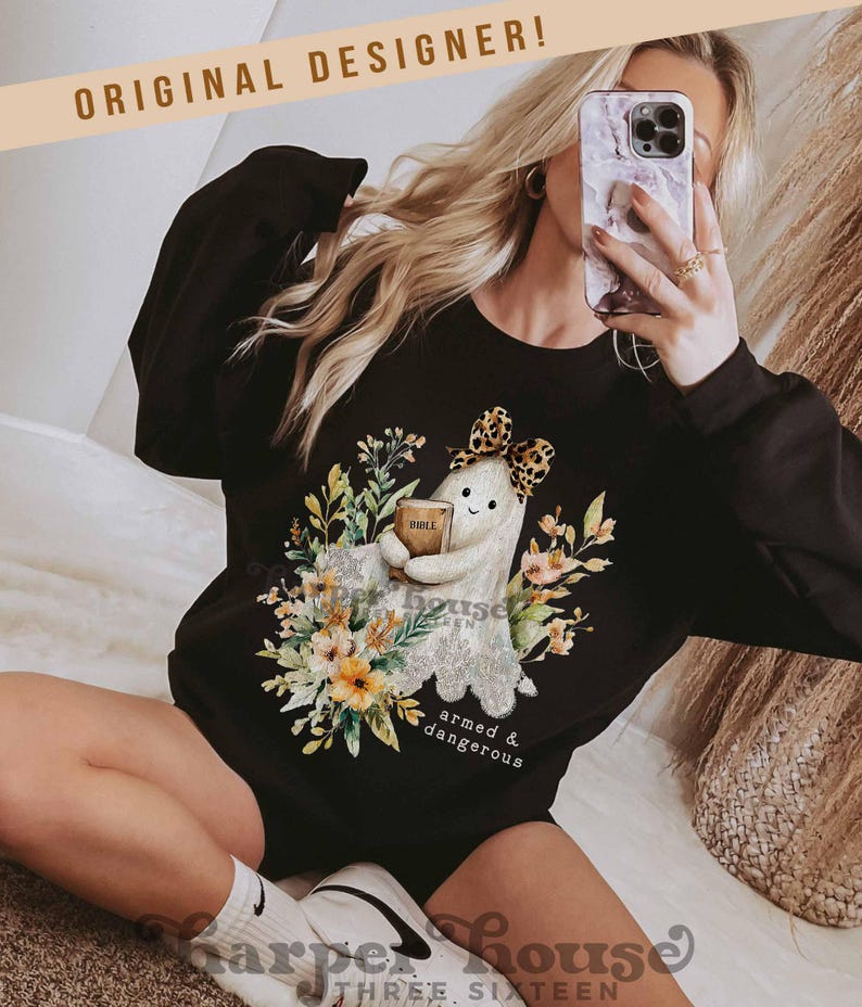 May include: Black sweatshirt featuring a ghost design holding a book labeled "Bible" surrounded by floral elements. The ghost wears a leopard print bow. Text on the sweatshirt reads "armed & dangerous". The words "Original Designer!" are at the top.