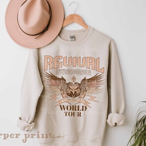 May include: A beige sweatshirt with a graphic print of an eagle with wings spread, a cross, and the text "REVIVAL WORLD TOUR". The text is in a vintage style font.