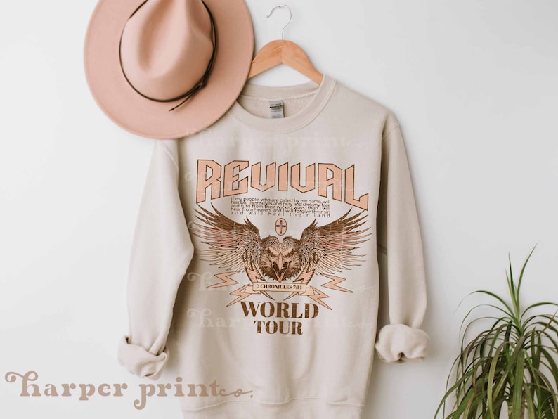 May include: A beige sweatshirt with a graphic print of an eagle with wings spread, a cross, and the text "REVIVAL WORLD TOUR". The text is in a vintage style font.
