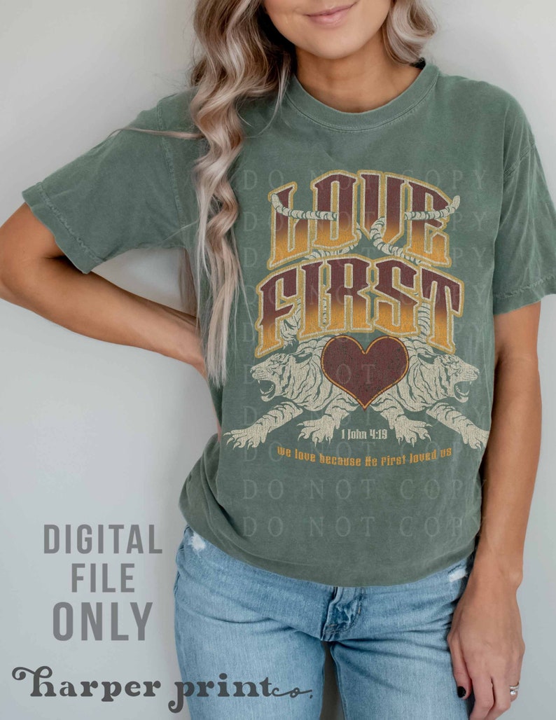 May include: A green t-shirt with a vintage-style graphic design featuring two tigers flanking a heart with the words "Love First" and the Bible verse "1 John 4:19"  "We love because He first loved us".