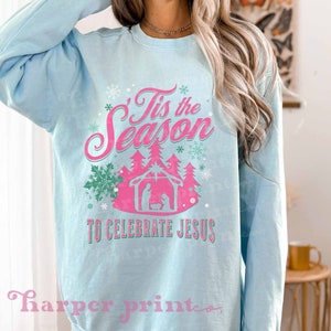 May include: A light blue sweatshirt with a pink and green design that says "Tis the Season to Celebrate Jesus". The design includes a manger scene with a star above it and snowflakes.