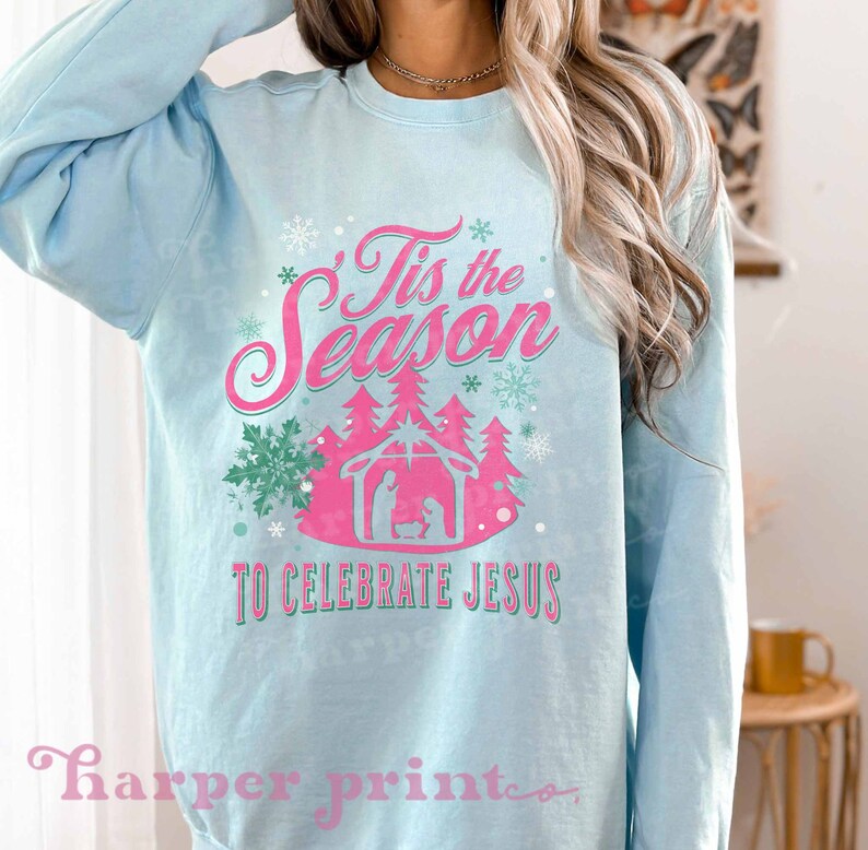 May include: A light blue sweatshirt with a pink and green design that says "Tis the Season to Celebrate Jesus". The design includes a manger scene with a star above it and snowflakes.