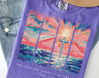 Puede incluir: Una camiseta morada con un gráfico de una cruz azul sobre olas del océano bajo un amanecer rosa y naranja. El texto dice "THE CROSS HAS SPOKEN, I AM FORGIVEN 1 Peter 2:24" y "harper house THREE SIXTEEN" está impreso debajo.