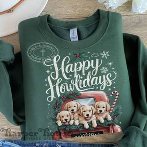 Op de afbeelding: Groen sweatshirt met een kerstontwerp met een rode vrachtwagen met vier golden retriever puppy's op de laadbak. De tekst "Happy Howlidays" is in wit op de voorkant van het sweatshirt gedrukt.