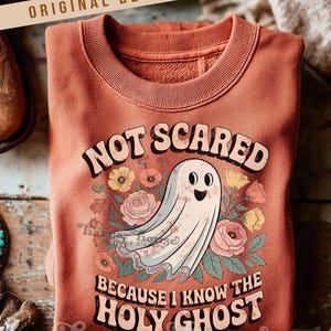 May include: A terracotta-colored sweatshirt with the text "NOT SCARED BECAUSE I KNOW THE HOLY GHOST" and a cartoon ghost illustration surrounded by flowers. The sweatshirt has a crew neck and long sleeves.