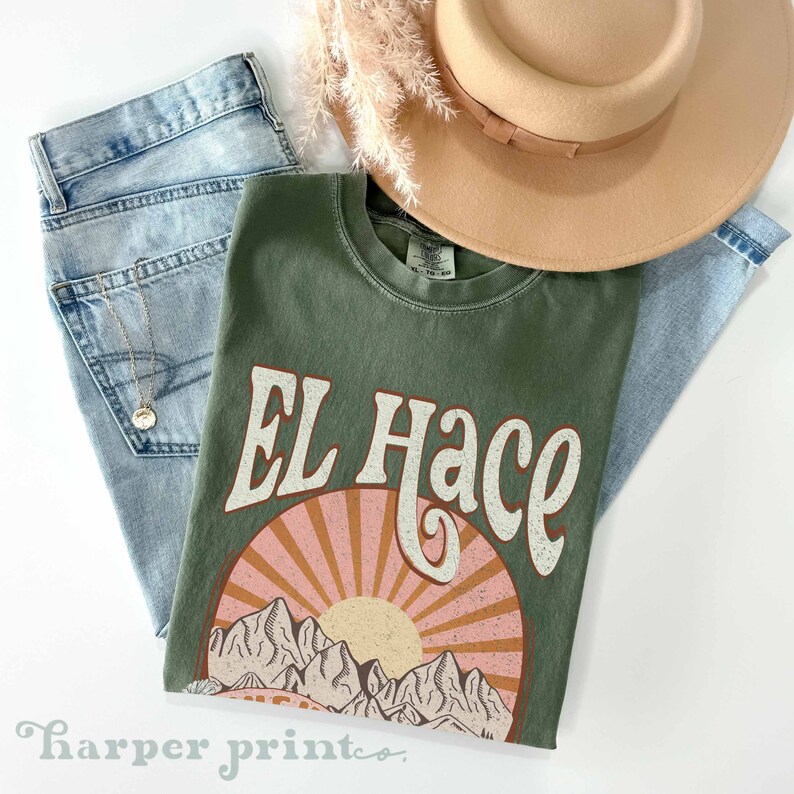 May include: A green t-shirt with a vintage-style graphic design featuring the text "EL Hace" and a sunset over mountains. The graphic is in shades of pink, orange, and brown.