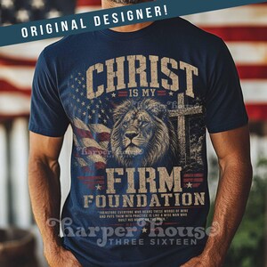 May include: Navy blue t-shirt with a graphic featuring a lion, cross, and American flag. The text reads "CHRIST IS MY FIRM FOUNDATION" and includes the phrase "THREE SIXTEEN". The shirt has a distressed, vintage look.