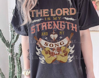 May include: A black short-sleeve t-shirt featuring the text THE LORD IS MY STRENGTH AND MY SONG. The graphic includes two acoustic guitars, a central heart with a dagger, roses, and lightning bolt accents in shades of orange, white, and tan.