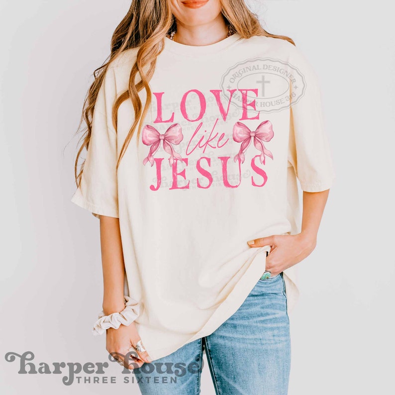 K&ouml;nnte beinhalten: Cremefarbenes T-Shirt mit dem Text "LOVE like JESUS" in Pink, mit zwei rosa Schleifen-Grafiken. Das Shirt hat eine lockere Passform und wird mit blauen Jeans getragen. Das Design beinhaltet ein Kreuz und die Worte "ORIGINAL DESIGNER" in einem Kreis.