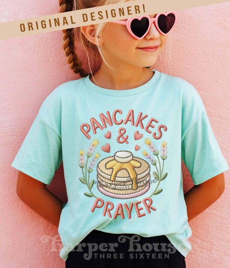 May include: A light blue t-shirt with the words "PANCAKES & PRAYER" embroidered in a reddish-brown color. The design includes a stack of pancakes with syrup, hearts, and floral accents. The text "ORIGINAL DESIGNER!" is above the shirt.