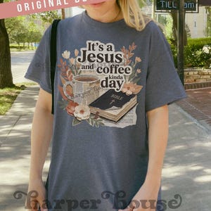 May include: A dark gray t-shirt with the text "It's a Jesus and coffee kinda day" and a graphic of a coffee cup, flowers, and a Bible. The shirt has short sleeves and a relaxed fit. The words "ORIGINAL DESIGNER!" are printed on a pink banner.