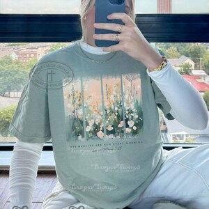 May include: A light green t-shirt with a floral design and the text "His mercies are new every morning" and a small text "Lamentations 3:23". The shirt has a distressed look and is made of a soft, comfortable material.